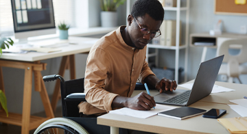 A person sitting on a desk, taking notes and using the computer.