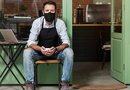 A maksed businessman holding a cup and sitting outside his shop with a table and lap top computer by his side.