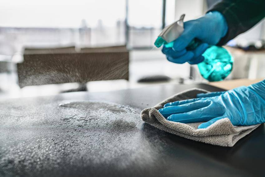 Image shows a pair of hands in blue rubber gloves using a disenfectant spray bottle and cleaning rag on a business office surface.