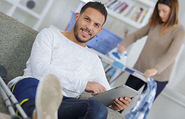 Man with injured leg sitting at home with table computer while his wife standing in the backgorund at an ironing board