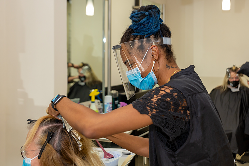 An African American woman wearing a face shield serving a customer at a hair salon.