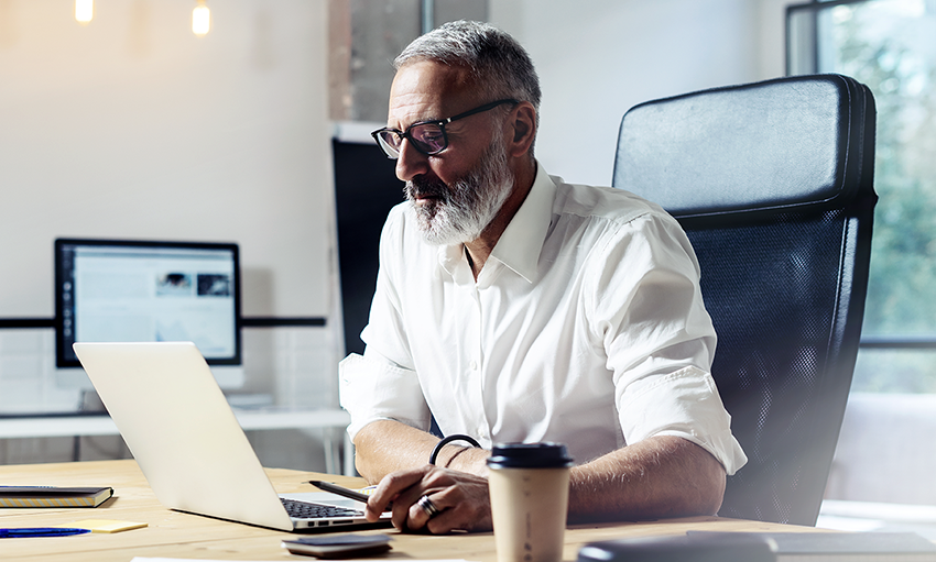 Elderly businessman with gray hair and beard, looking intently at the screen as he enters information on a laptop computer 