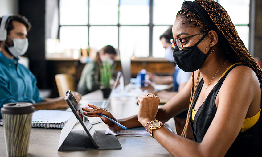 An African American woamn in foreground and a group of co-workers wearing face masks sit opposite each other at a workstation table 