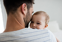 A father viewed from behind holds a baby facing the camera over the father's shoulder.