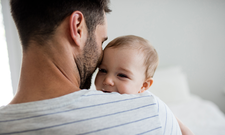 A father viewed from behind holds a baby facing the camera over the father's shoulder.