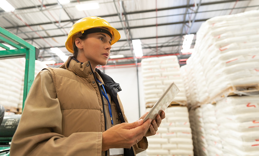 A woman in hardhat and work jacket uses a computer tablet during a warehouse survey for safety risks