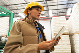 A woman in hardhat and work jacket uses a computer tablet during a warehouse survey for safety risks